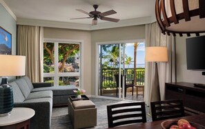 A hotel room with a sofa, ottoman, dining table, chairs, a TV, and a balcony with a view of palm trees and a building. - ALOHA!! GRAND OCEAN VIEW  1-bedroom Villa with AC in Kaanapal, Steps to Beach! (Lahaina)