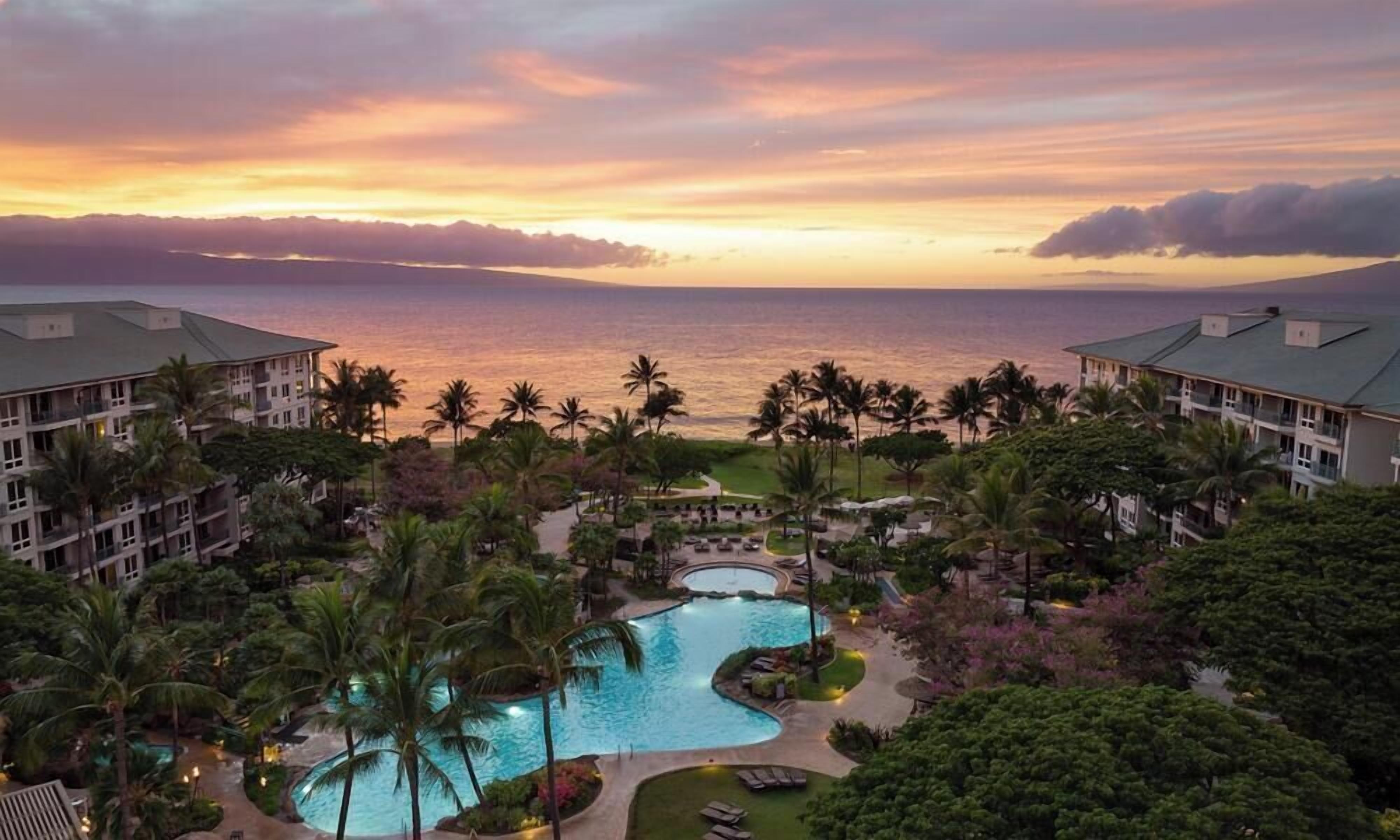 A resort with a swimming pool, palm trees, and a view of the ocean at sunset.