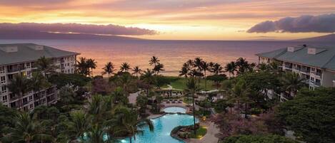 A resort with a swimming pool, palm trees, and a view of the ocean at sunset.