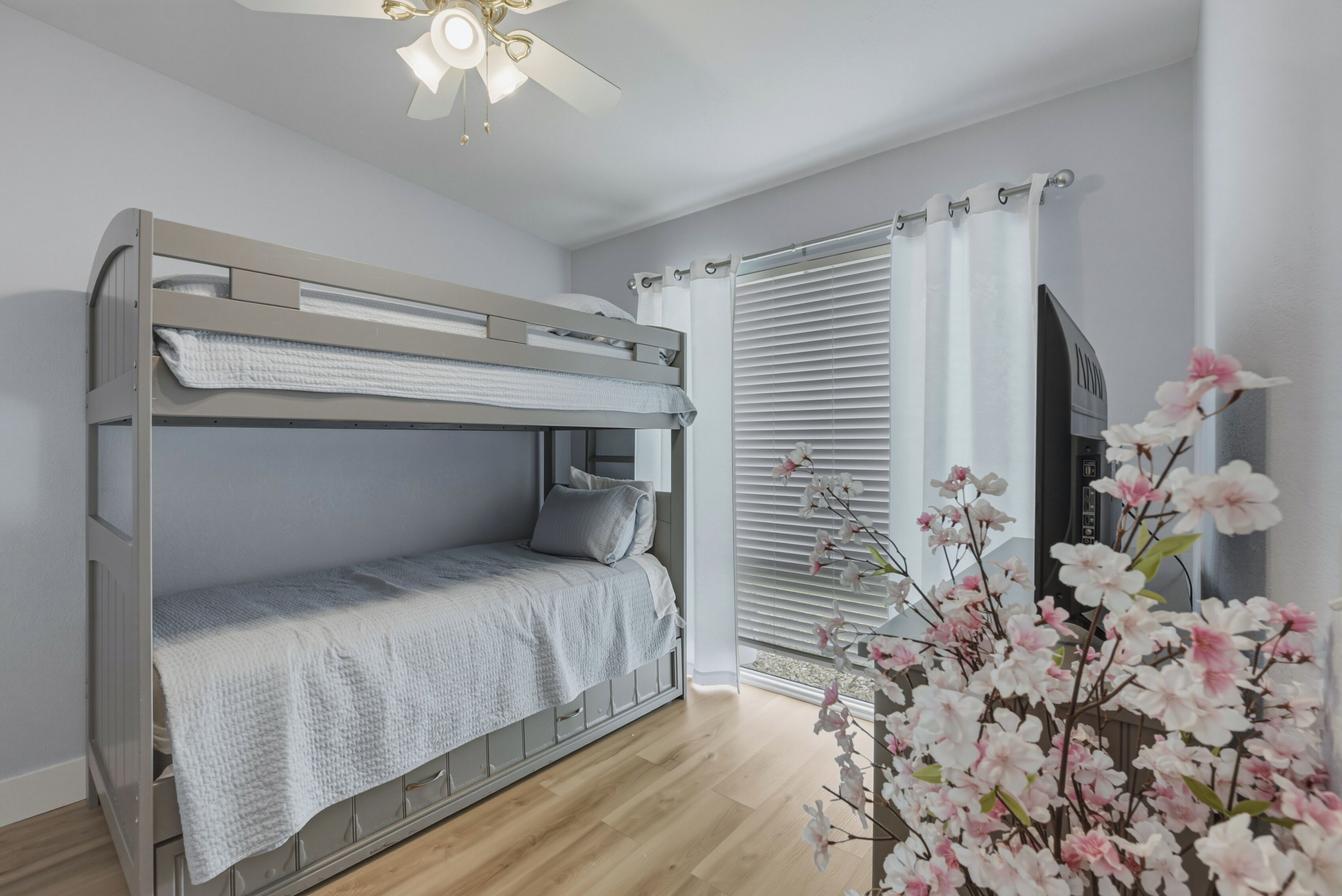 A bunk bed with a ceiling fan, a window with blinds, and a flower arrangement.