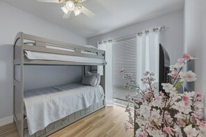 Een stapelbed met een plafondventilator, een raam met zonwering en een bloemstuk.