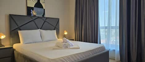 Una habitación de hotel con una cama grande, un cabecero, dos lámparas de noche, una ventana con cortinas y una obra de arte colgada en la pared.