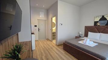 A hotel room with a bed, bedside table, a flat-screen TV, a potted plant, and a wooden floor.