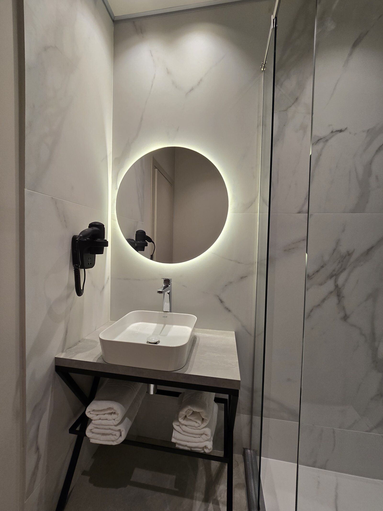 Un bany modern amb una paret de marbre, un mirall rodó amb llum, un lavabo blanc i un suport per a tovalloles.