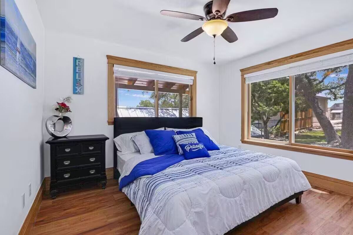 A bedroom with a bed, a nightstand, a ceiling fan, and large windows.