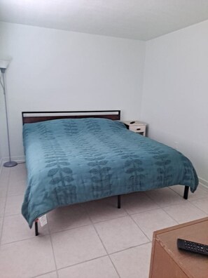 Un lit simple avec une tête de lit, une table de chevet et une lampe sur pied, dans une pièce aux murs unis et au sol carrelé.