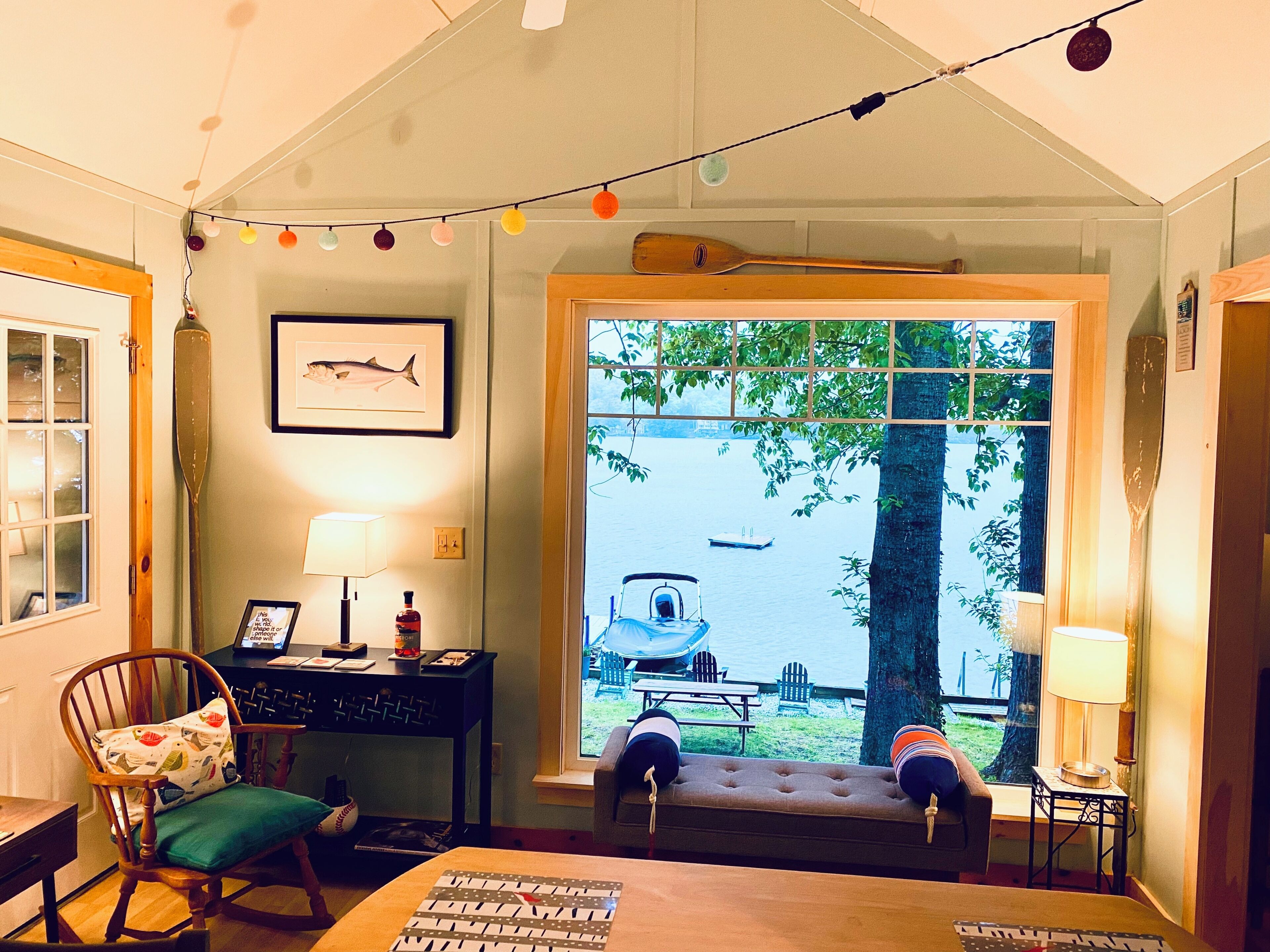 A cozy room with a view of a lake, a wooden paddle, a framed fish picture, and a desk with a lamp.