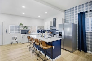 A modern kitchen with a central island, bar stools, a stainless steel refrigerator, and a blue accent wall. - The Victory Lap | Hot Tub • Fire Pit • Game Lounge (Indianapolis)