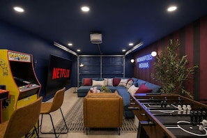 Een kamer met een grote flatscreen televisie waarop 'NETFLIX' staat, een tafelvoetbaltafel, een bank en een aan de muur bevestigde projector.