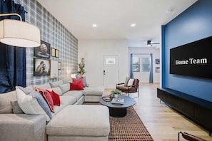 A modern living room with a sectional sofa, a coffee table, a TV wall unit, and a ceiling fan. - The Victory Lap | Hot Tub • Fire Pit • Game Lounge (Indianapolis)