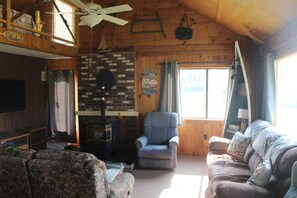 A cozy cabin interior with a fireplace, a television, a sofa, and a window with curtains. - Mascoma Lake Quiet Cottage - Lakefront living! (Lebanon)