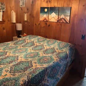 Ein Schlafzimmer mit Holzwand, einem Bett mit gemustertem Überwurf, einem Nachttisch, einer Lampe und einem Wandbild von Bergen.