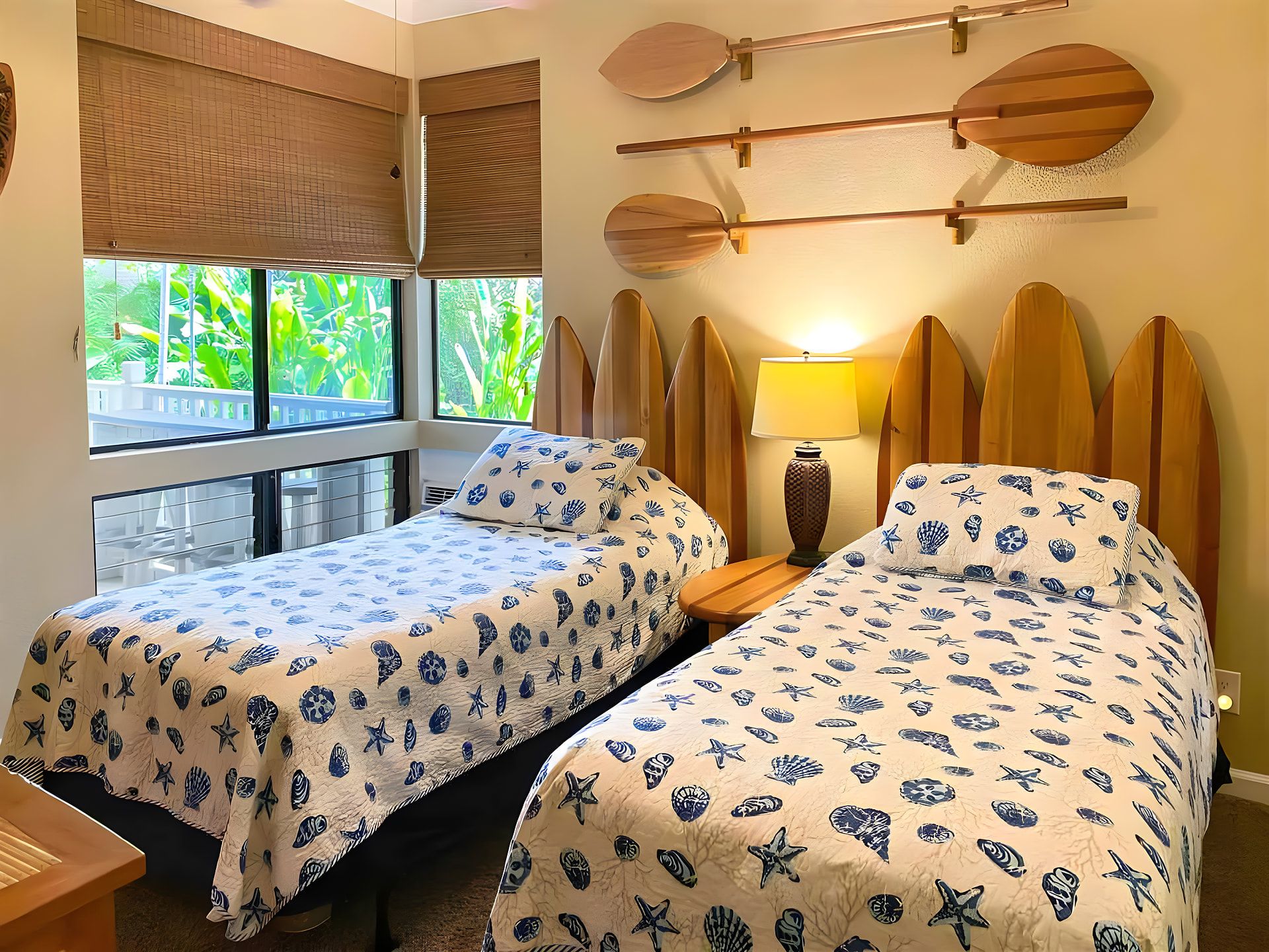 A room with two beds, a window with a view of greenery, and surfboards mounted on the wall.
