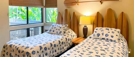A room with two beds, a window with a view of greenery, and surfboards mounted on the wall.