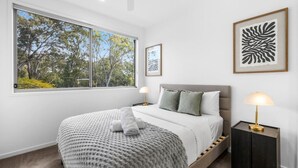 A bedroom with a large window, a bed with a grey headboard, two bedside tables with lamps, and framed artwork on the walls.