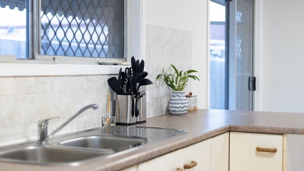 A kitchen with a double sink, a window with a metal screen, a potted plant, and a utensil holder. - Heart of Sunnybank | Prime Location | Great Value (Sunnybank)