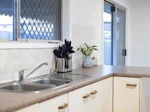 Una cocina con un lavamanos doble, una ventana con mosquitero, una planta en maceta y un recipiente para utensilios.