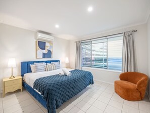 A bedroom with a bed, nightstand, chair, and a large window with curtains. - Heart of Sunnybank | Prime Location | Great Value (Sunnybank)