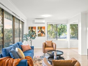 Una sala de estar moderna con un sofá azul, un sillón de cuero marrón y una mesa de centro. Hay ventanas amplias con vistas a zonas verdes.