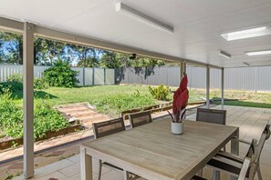 An outdoor patio with a wooden table and chairs, a potted plant, and a view of a backyard with a fence and greenery.