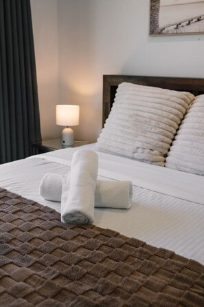 A neatly made bed with white linens, a brown textured blanket, and two rolled towels.