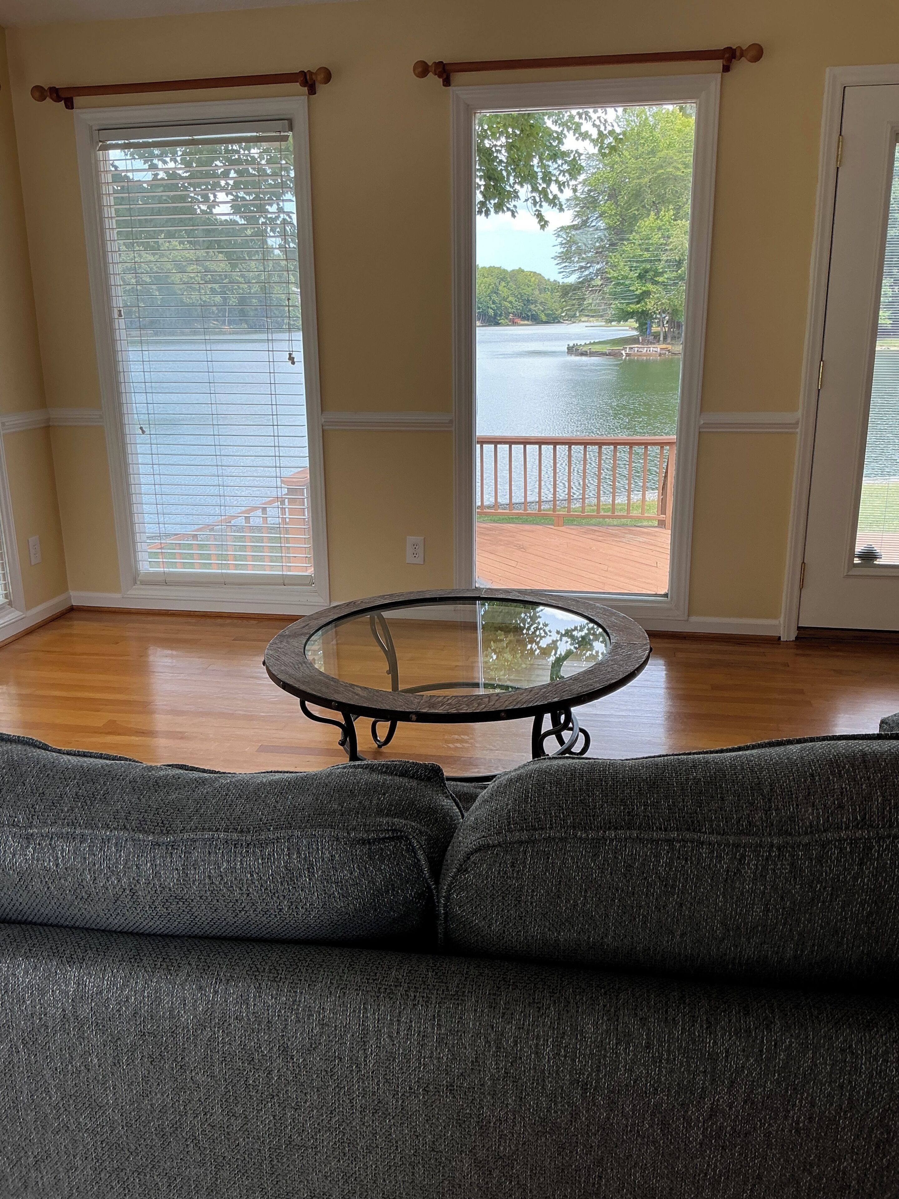 A room with a round glass table, a gray sofa, and large windows with a view of a lake and deck.