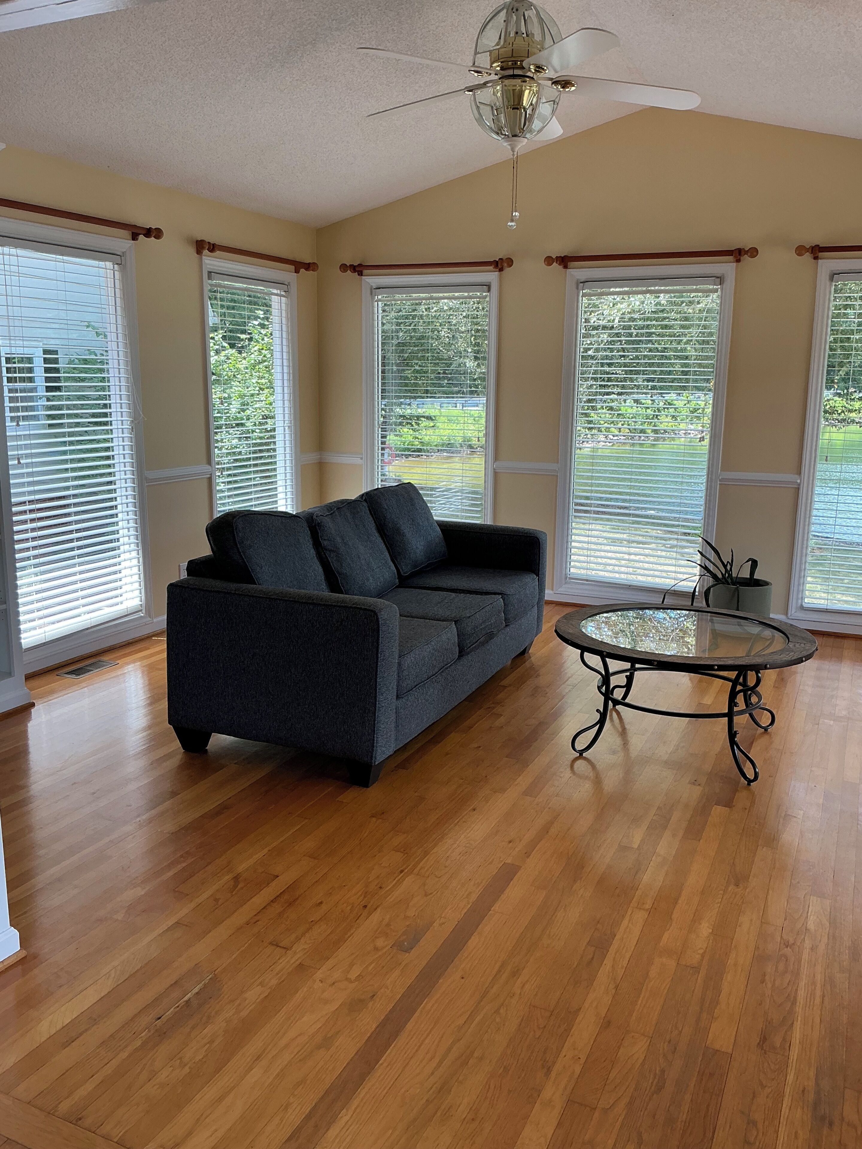 A room with a dark gray sofa, a glass-top coffee table, wooden flooring, and large windows with blinds.