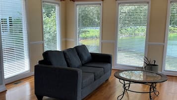 A room with a dark gray sofa, a glass-top coffee table, wooden flooring, and large windows with blinds.