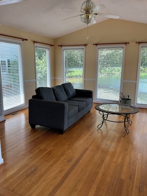A room with a dark gray sofa, a glass-top coffee table, wooden flooring, and large windows with blinds. - Simple Lake house in charming Summerfield with exceptional lake views! (Summerfield)