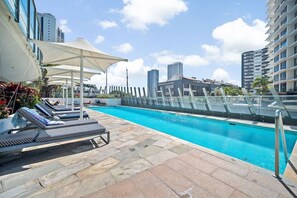 Ein Pool auf dem Dach mit Liegestühlen und Sonnenschirmen, umgeben von Hochhäusern.