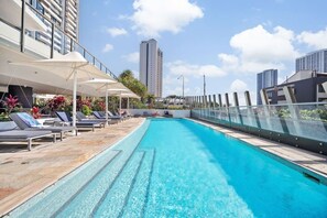 Une piscine sur le toit, agrémentée de chaises longues et de parasols, et entourée d’immeubles de grande hauteur.