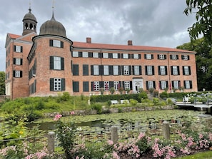 Um edifício grande de tijolos com uma cúpula central e duas cúpulas menores, cercado por um lago com vitórias-régias e flores cor-de-rosa.