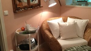 Um quarto acolhedor com teto inclinado, um sofá de vime, uma mesa redonda pequena com uma luminária e uma janela com vista para árvores.