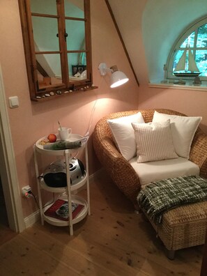 Ein gemütliches Zimmer mit schrägen Wänden, einem Korbssessel, einem kleinen runden Tisch mit Lampe und einem Fenster mit Blick auf Bäume.