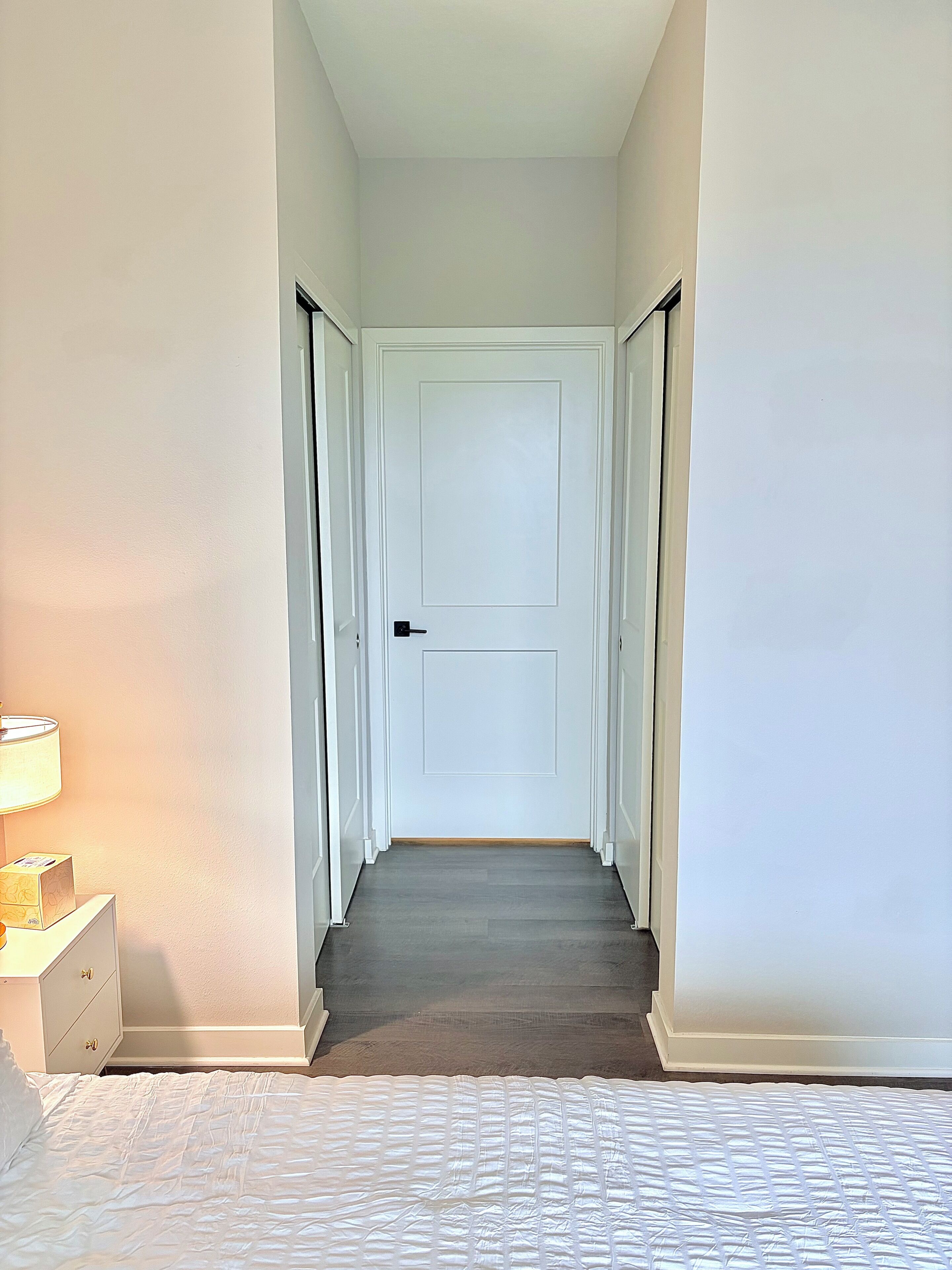 A hallway with a closed door, a bedside table with a lamp, and a bed with a white quilt.