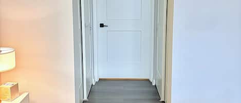 A hallway with a closed door, a bedside table with a lamp, and a bed with a white quilt.