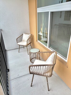 Un balcone con due sedie in vimini e un piccolo tavolino rotondo, una finestra che si affaccia sulla strada e un muro con una superficie strutturata.
