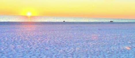 Una playa con atardecer, mar tranquilo y huellas en la arena.