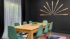 En matplats med ett träbord, gröna stolar, en svart vägg med en dekoration i form av en fladdermus och ett fönster med tunna, genomskinliga gardiner.