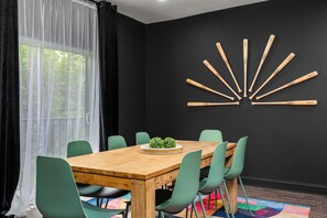 A dining area with a wooden table, green chairs, a black wall with a bat decoration, and a window with sheer curtains. - The Stables 2 - Minigolf, Firepits, Games, BBQ (Louisville)