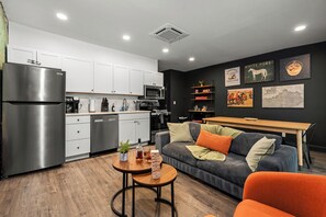 A modern kitchen with white cabinets, a stainless steel refrigerator, and a microwave. There is a dining area with a wooden table and a sofa. The walls are decorated with framed artwork. - The Stables 2 - Minigolf, Firepits, Games, BBQ (Louisville)