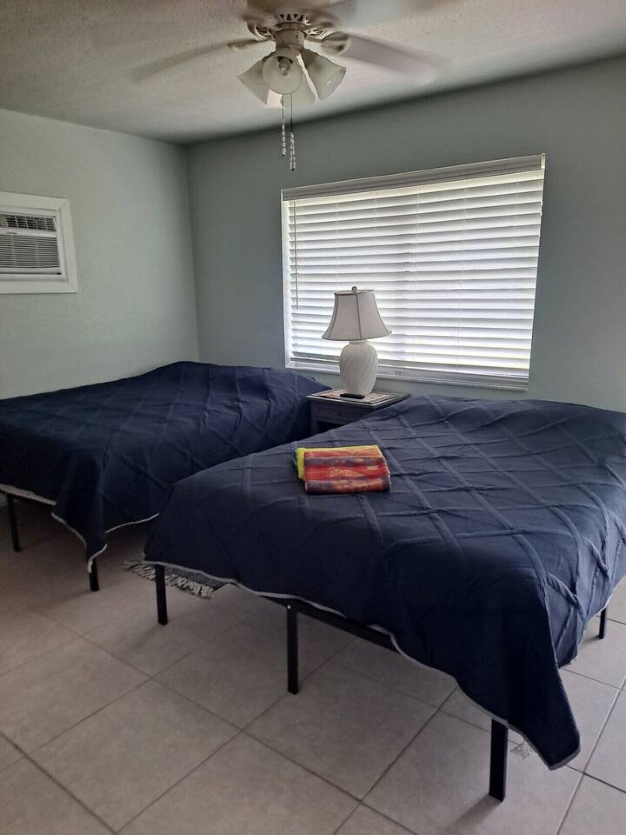 A room with two beds, a ceiling fan, a window with blinds, and a bedside table with a lamp.