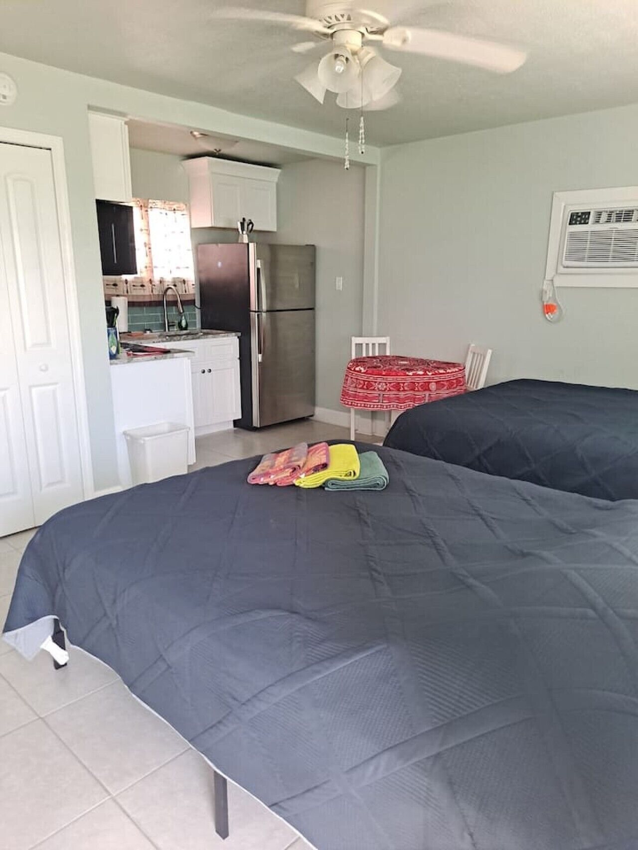 Un dormitorio con dos camas, una kitchenette, un ventilador de techo y una pequeña mesa con una silla.