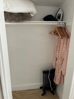 A closet with a hanging rack, a radiator, and a shelf with an iron and a pillow.