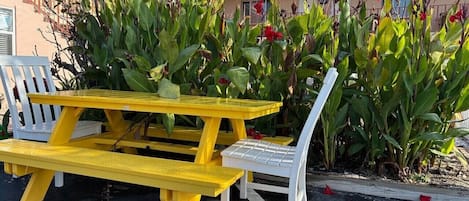 Une table de pique-nique jaune et un banc blanc se trouvent sur un parking, avec des fleurs rouges et un feuillage vert en arrière-plan.