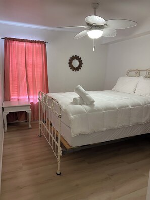 Ein Schlafzimmer mit einem Bett, einem Nachttisch, einem weißen Deckenventilator und einem Fenster mit rosa Vorhängen.
