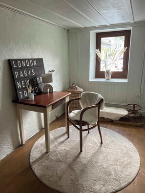 Une pièce avec un tapis rond, une table en bois, une chaise, un luminaire fixé au mur, une fenêtre donnant sur un paysage agréable et un panneau décoratif.