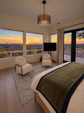Ein Schlafzimmer mit einem großen Bett, zwei Stühlen, einem Fernseher und Blick auf die Wüste bei Sonnenuntergang.