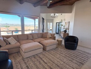 Ein Wohnzimmer mit einer Eckcouch, einem Hocker und einem schwarzen Sessel. Ein großes Fenster bietet einen Ausblick ins Freie.