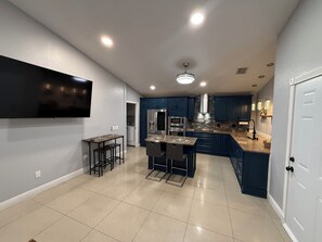 Cozinha moderna com armários azul escuro, uma ilha central, eletrodomésticos de aço inoxidável e uma TV de tela plana na parede.
